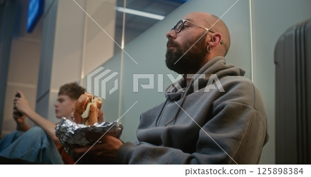 Airport Terminal: Man Eating Fast Food Burger while Sitting on the Floor, Waiting for Delayed Flight 125898384