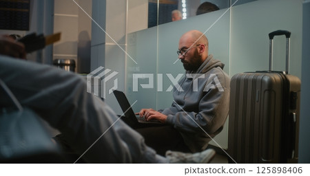 Airport Terminal: Man Doing Remote Work on Laptop, Sitting on the Floor in Airline Hub Lounge Airport Terminal: Man Doing Remote Work on Laptop, Sitting on the Floor in Airline Hub Lounge 125898406