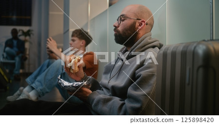 Man Eating Fast Food Burger while Sitting on the Floor in Departure Lounge of Airline Hub Man Eating Fast Food Burger while Sitting on the Floor in Departure Lounge of Airline Hub 125898420