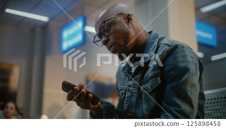 Man Eating Burger and Using Smartphone Sitting in Boarding Lounge of Airport Terminal 125898458