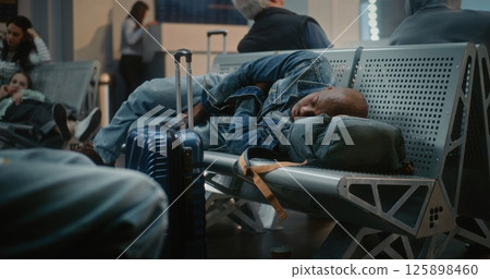 Tired Man with Suitcase and Backpack Sleeping on the Seats in Departure Lounge of Airport Terminal Tired Man with Suitcase and Backpack Sleeping on the Seats in Departure Lounge of Airport Terminal 125898460