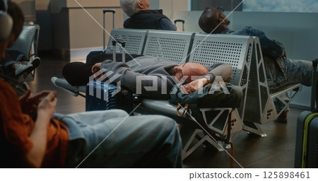 Exhausted Man with Luggage Sleeping on the Seats in Boarding Lounge of Airline Hub Exhausted Man with Luggage Sleeping on the Seats in Boarding Lounge of Airline Hub 125898461