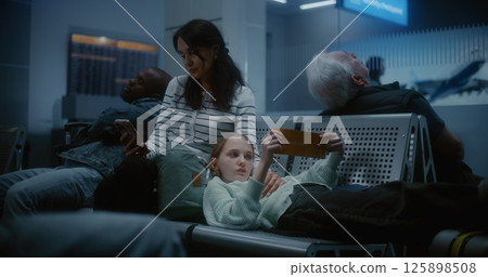 Boarding Lounge of Airline Hub at Night: Tired Teen Girl Lying on Seats, Woman Using Smartphone 125898508