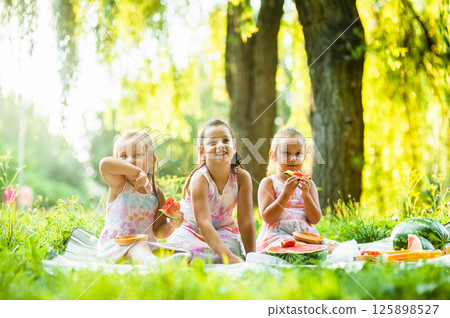 The sisters are sitting together enjoying a delicious watermelon in the open air. The sisters are sitting together enjoying a delicious watermelon in the open air. 125898527