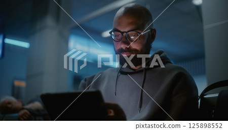 Tired and Sleepy Man Using Tablet Computer, Sitting in Departure Lounge of Airline Hub at Night Tired and Sleepy Man Using Tablet Computer, Sitting in Departure Lounge of Airline Hub at Night 125898552
