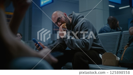 Departure Lounge of Airline Hub at Night: Man Eating Burger and Scrolling Phone 125898558