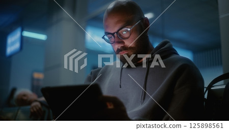 Airport Terminal at Night: Tired and Sleepy Man Yawning, Falling Asleep When Using Digital Tablet Airport Terminal at Night: Tired and Sleepy Man Yawning, Falling Asleep When Using Digital Tablet 125898561
