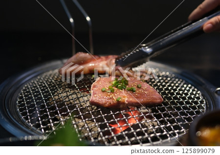 Close-up of hands masterfully flipping meats on a smoky grill, set against a blurred natural backdrop 125899079