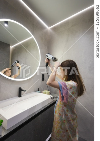 Teen girl in a floral robe dries her hair in front of a mirror with a hairdryer in a modern bathroom Teen girl in a floral robe dries her hair in front of a mirror with a hairdryer in a modern bathroom 125899662