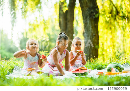 The sisters are sharing a picnic and laughing while eating watermelon. Their dresses match perfectly, making them look even more alike. The sisters are sharing a picnic and laughing while eating watermelon. Their dresses match perfectly, making them look even more alike. 125900059