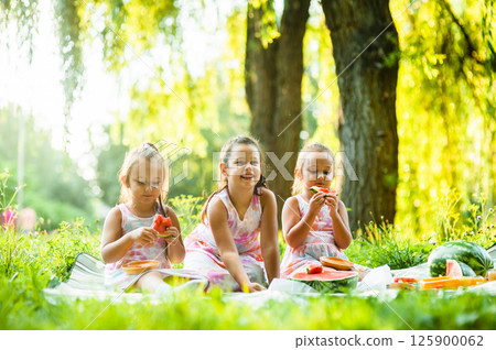 The three sisters are having a picnic surrounded by green grass and bright sunshine. The little twins share playful glances as they eat their watermelon, their dresses perfectly matching. 125900062
