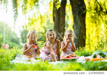 The sisters are sitting on a picnic blanket, enjoying a beautiful summer day. The twins giggle as they take big bites of watermelon, their matching dresses making them look even cuter. 125900065