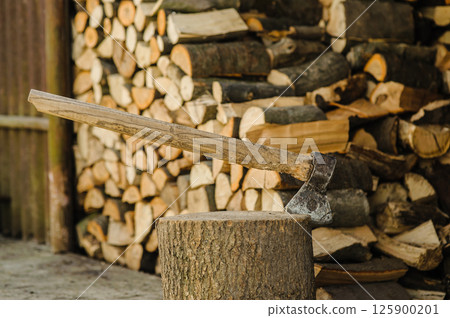 An axe is stuck in a wooden log, standing firmly in place. In the background, there are chopped and neatly stacked firewood. An axe is stuck in a wooden log, standing firmly in place. In the background, there are chopped and neatly stacked firewood. 125900201