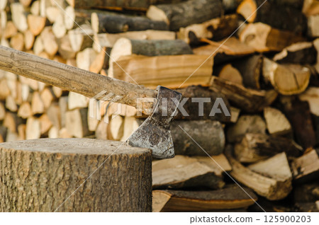 A sharp axe is embedded in a sturdy log. In the background, there are stacked firewood pieces.  125900203