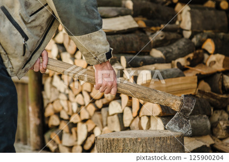 A firm hand holds an axe embedded in a stump. Stacks of chopped wood rest in the background. 125900204