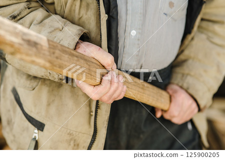 An old man's hand clutches an axe. Behind it are stacks of firewood.  125900205