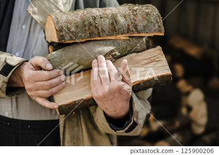 Rough, veined hands carry freshly chopped logs. Behind them, firewood is piled high. Rough, veined hands carry freshly chopped logs. Behind them, firewood is piled high. 125900206