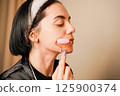 Using a facial roller, a woman massages her jawline. A soothing skincare routine. Her natural beauty and elegant earrings complete the sophisticated scene. The lighting creates a peaceful mood. 125900374