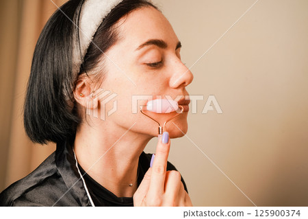 Using a facial roller, a woman massages her jawline. A soothing skincare routine. Her natural beauty and elegant earrings complete the sophisticated scene. The lighting creates a peaceful mood. Using a facial roller, a woman massages her jawline. A soothing skincare routine. Her natural beauty and elegant earrings complete the sophisticated scene. The lighting creates a peaceful mood. 125900374