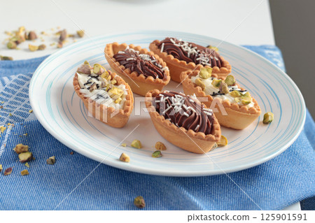 Sand dough tartlets with chocolate filling, pistachio nuts on white plate. Dessert for party, holiday.  125901591