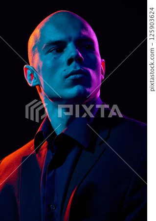 Close-up portrait of handsome bald man in dark shirt and jacket,posing confidently on dark background in multi-colored neon light Close-up portrait of handsome bald man in dark shirt and jacket,posing confidently on dark background in multi-colored neon light 125903624