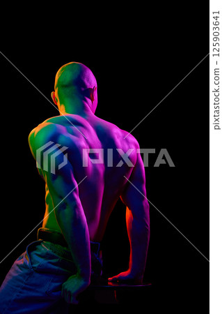 Back view of young bald man, leaning on his hands, posing shirtless, showing muscular body on dark background in multi-colored neon light. Back view of young bald man, leaning on his hands, posing shirtless, showing muscular body on dark background in multi-colored neon light. 125903641