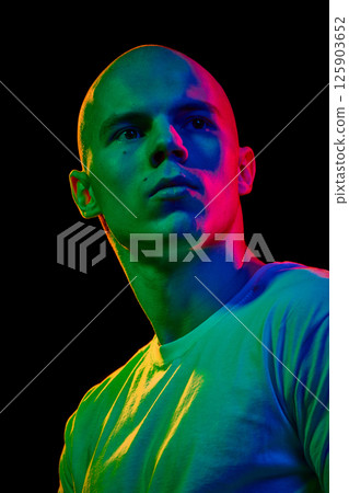 Close-up portrait of young confident bald man in white t-shirt, looking upwards with serious expression on dark background 125903652