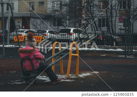 A child sits alone on a swing in an empty playground. A little girl in a pink winter coat sits alone on a swing in an empty playground in winter, with cars parked behind a fence in the background. A child sits alone on a swing in an empty playground. A little girl in a pink winter coat sits alone on a swing in an empty playground in winter, with cars parked behind a fence in the background. 125903835