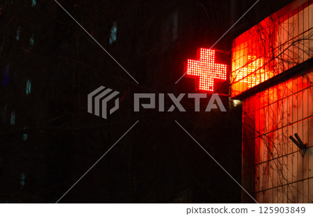 A bright red cross made of small bulbs glows brightly against a dark background, indicating the presence of a hospital or medical facility at night. Red cross glows in the dark on a house. 125903849