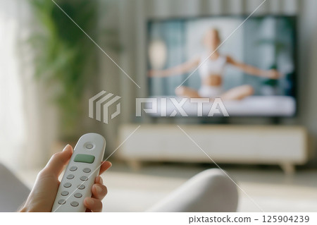 A close-up of a hand holding a remote control, focusing on a yoga instructor displayed on a TV in a modern and serene home setting, emphasizing fitness and relaxation. Blurred background 125904239
