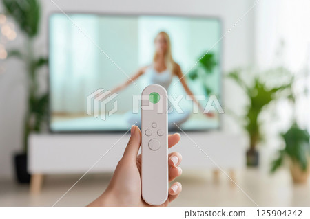 A close-up of a hand holding a remote control, focusing on a yoga instructor displayed on a TV in a modern and serene home setting, emphasizing fitness and relaxation. Blurred background 125904242