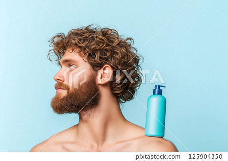 Bearded man with wavy hair holding a blue shampoo bottle on his shoulder. The light background and clear focus highlight personal grooming and care in an aesthetic composition. Men's cosmetics. 125904350