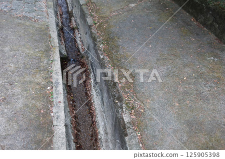 a narrow concrete drainage channel running through a paved area. The channel has a shallow flow of water and is flanked by worn concrete surfaces. The surrounding pavement is weathered with copyspace a narrow concrete drainage channel running through a paved area. The channel has a shallow flow of water and is flanked by worn concrete surfaces. The surrounding pavement is weathered with copyspace 125905398