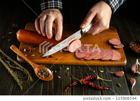 A person skillfully slices sausage on a wooden cutting board, surrounded by herbs, spices, and fresh garlic in a warm, inviting kitchen atmosphere 125906594