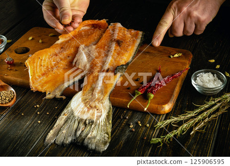 Hands season a fresh salmon fillet with spices on a wooden cutting board. Various herbs, salt, and a red chili pepper add vibrant color and flavor in a cozy kitchen atmosphere 125906595
