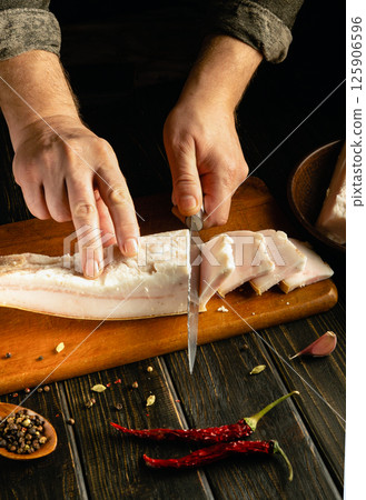 A chef focuses intently as he expertly slices fresh fish on a wooden cutting board. Various spices and red chili peppers are arranged nearby, enhancing the culinary atmosphere 125906596
