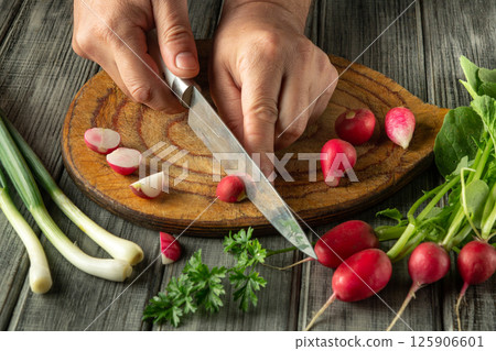 Hands skillfully slice vibrant radishes on a wooden board, surrounded by fresh greens and herbs, capturing the essence of healthy cooking in a warm kitchen atmosphere 125906601