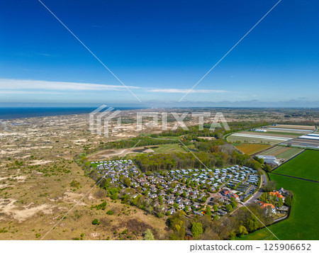 A stunning aerial view of a coastal town with red-tiled rooftops, neatly aligned houses, and a vast sandy beach stretching into the distance under a clear blue sky. 125906652