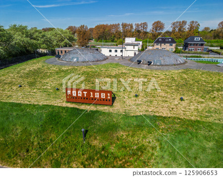 Aerial view of a historic fort with round domes, green grass-covered walls, walking paths, and adjacent buildings, surrounded by trees and open parkland under bright daylight. Aerial view of a historic fort with round domes, green grass-covered walls, walking paths, and adjacent buildings, surrounded by trees and open parkland under bright daylight. 125906653