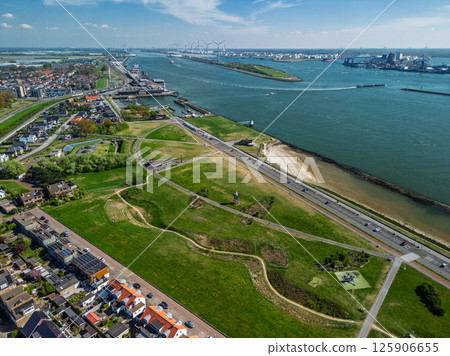 A coastal view showing a quiet seaside town with a rocky shoreline, green open fields, a road with passing vehicles, and residential buildings in the distance under a clear blue sky. 125906655
