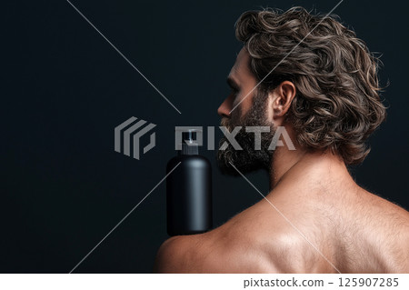 Man with curly hair holding a shampoo bottle against a dark background. Curly haired man with beard with a shampoo bottle standing on his shoulder, highlighting hair care concepts. 125907285