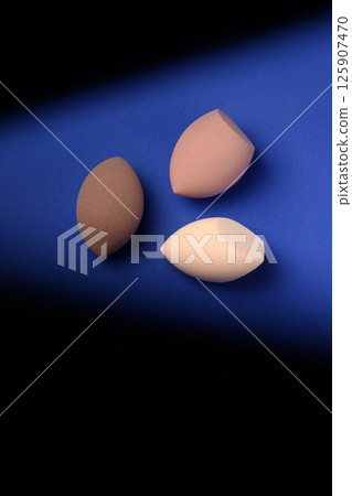 Three different cosmetic blenders on a blue background with a contrast shadow. Brown makeup sponges with pointed cutouts. Place for text. creative promotional photo for makeup artists. Three different cosmetic blenders on a blue background with a contrast shadow. Brown makeup sponges with pointed cutouts. Place for text. creative promotional photo for makeup artists. 125907470