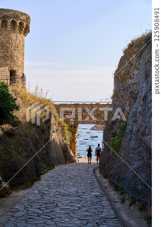 A scenic pathway in Tossa de Mar leads toward the sea, framed by stone walls and a historic tower, with two people walking. A scenic pathway in Tossa de Mar leads toward the sea, framed by stone walls and a historic tower, with two people walking. 125908491