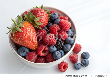 Bowl filled with fresh mixed berries, vibrant red strawberries, blueberries, and raspberries on a white table 125908883