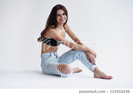 Charming skinny beautiful girl with perfect body in jeans and black top sits on white background Charming skinny beautiful girl with perfect body in jeans and black top sits on white background 125910246
