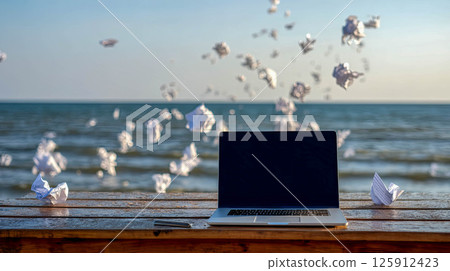 Creative workspace by the sea with a laptop and flying paper balls representing ideas concept 125912423