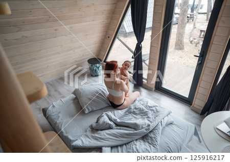 A red-haired Caucasian woman sits in bed with her little son in a country house with panoramic windows. 125916217