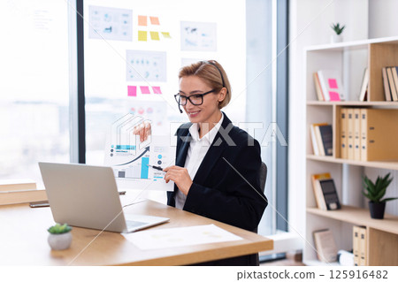Businesswoman in office delivers online presentation showing data charts and discussing trends. Female professional, mid-30s, engaged in analysis. Businesswoman in office delivers online presentation showing data charts and discussing trends. Female professional, mid-30s, engaged in analysis. 125916482