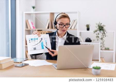 Businesswoman in office delivers online presentation showing data charts and discussing trends. Female professional, mid-30s, engaged in analysis. Businesswoman in office delivers online presentation showing data charts and discussing trends. Female professional, mid-30s, engaged in analysis. 125916483