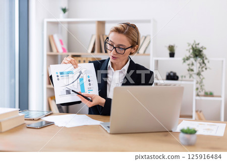 Confident businesswoman in formal outfit presenting financial documents at office desk with laptop. Office plant decor is visible in background Confident businesswoman in formal outfit presenting financial documents at office desk with laptop. Office plant decor is visible in background 125916484
